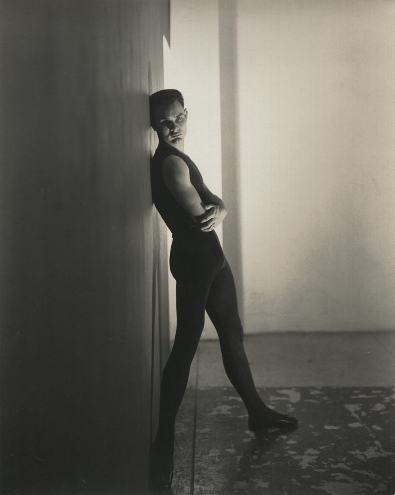 This is a black-and-white photograph of a man in a black t-shirt and black tights seen from the side leaning with his back against a wall.