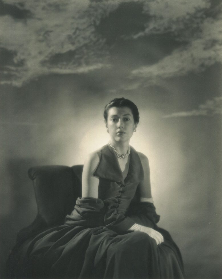 Fidelma Cadmus Kirstein sitting in a chair wearing a dark sleeveless top and shawl, pearl choker and white gloves