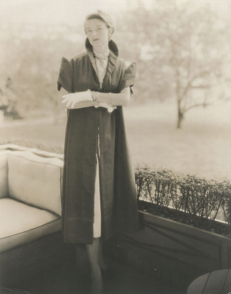 Woman standing wearing a long, dark, sleeveless coat over a light dress and white gloves