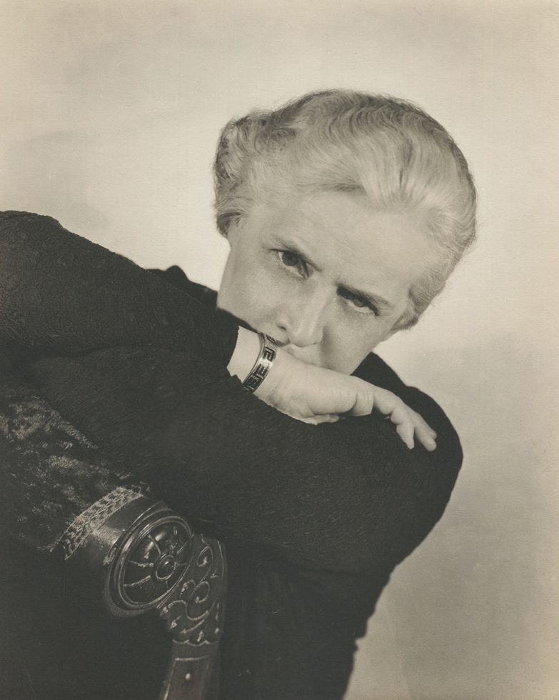 An older woman with white hair with her arms crossed over the back of a chair and her chin resting on her arms