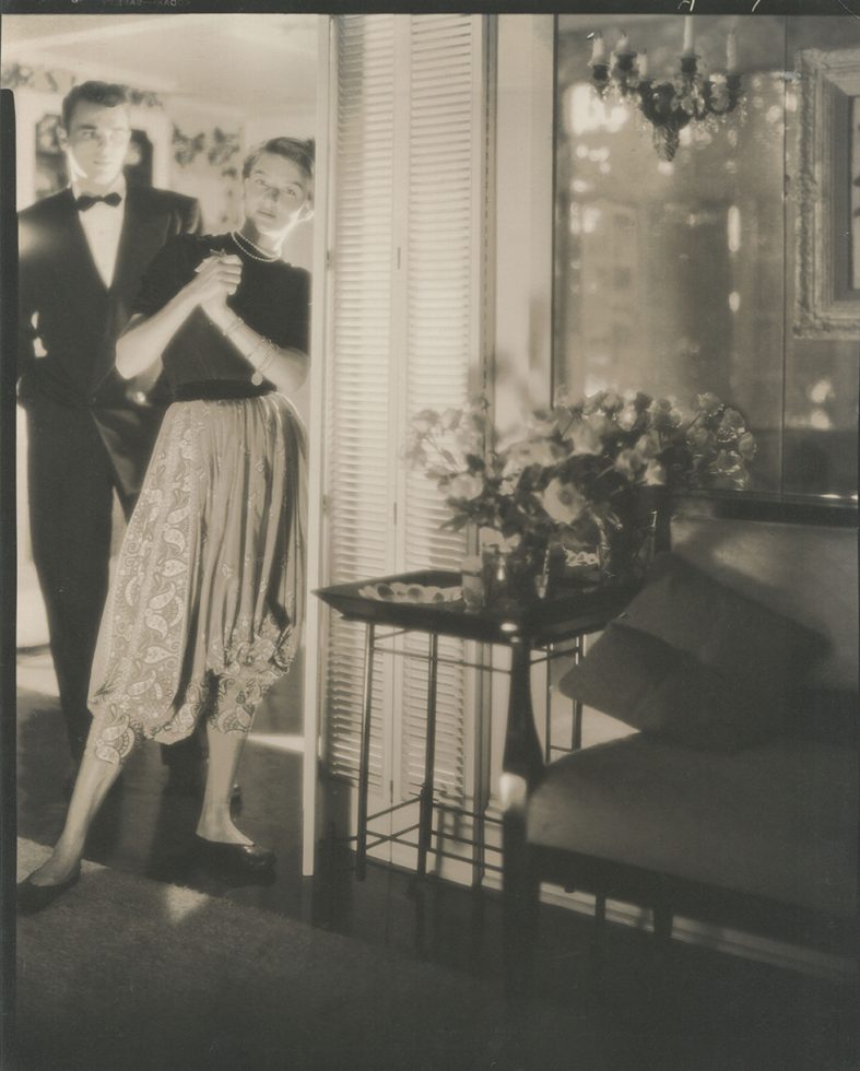 Young man and woman he is wearing a tuxedo and she is wearing paisley, loose fitting trousers, a dark top and a pearl necklace