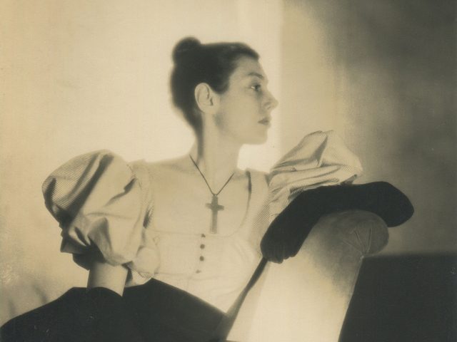 Sepia-toned photograph of Fidelma Cadmus Kirstein in profile with her hair in a bun wearing a large cross around her neck and long, black, velvet gloves.