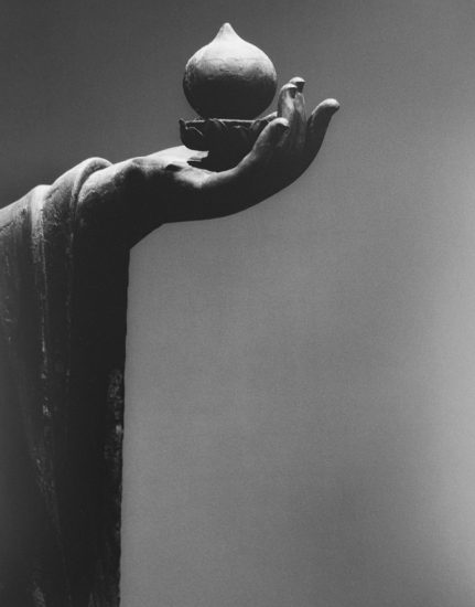 A sculpted hand holds a round object with a pointed top, balanced delicately on the fingertips against a smooth gray background.