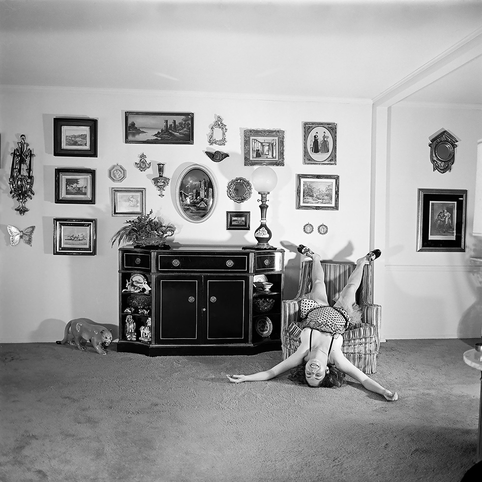 A black and white self portrait photo by Meryl Meisler of the artist upside down in an easy chair in a suburban living room in 1975.