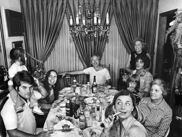 A black-and-white photograph by Meryl Meisler of three Long Island families gathered around a table for Rosh Hashana dinner in 1974.