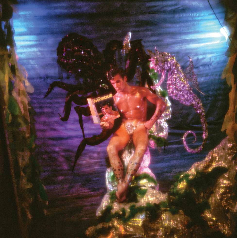 A photograph by James Bidgood of a man sitting underwater reading a magazine called "The Young Physique" next to a purple seahorse with a giant octopus in the background.