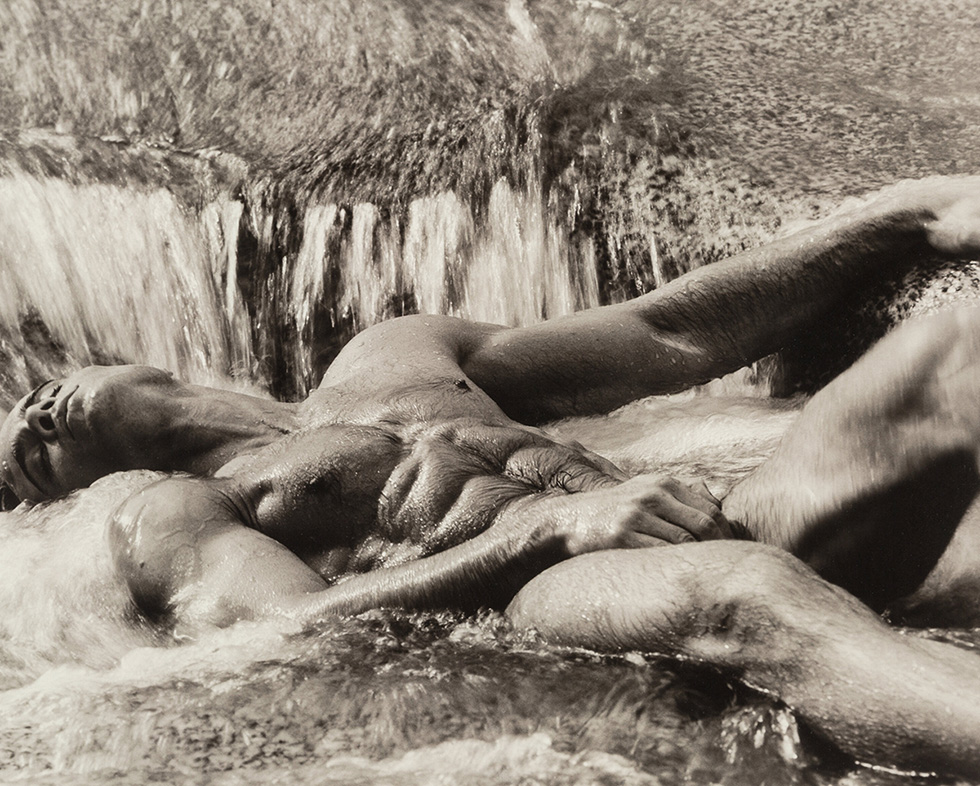 This is a sepia-toned black-and-white photograph of a naked man reclining in rushing water.