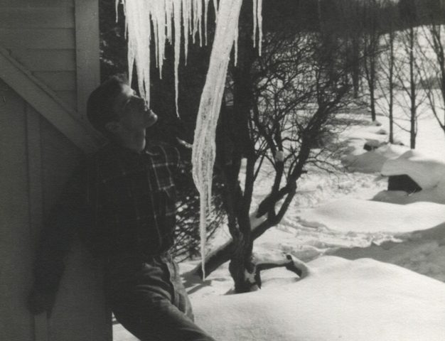 Photograph of a young man in a flannel shirt and wide-legged jeans standing in the snow against a house contemplating a row of icicles hanging from a gutter above his head.