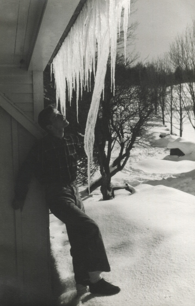 This is a black-and-white photograph of a young man in a flannel shirt and wide-legged jeans standing in the snow against a house contemplating a row of icicles hanging from a gutter above his head.