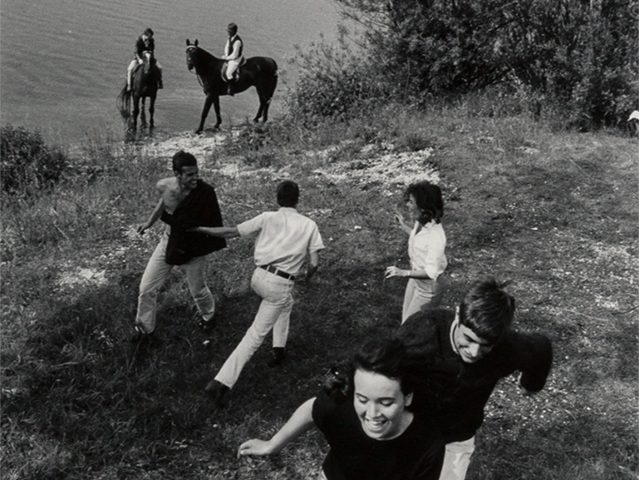 A black-and-white photo by Will McBride of five young adults frolicking on a hill near Munich with two others on horseback in the distance by a body of water.