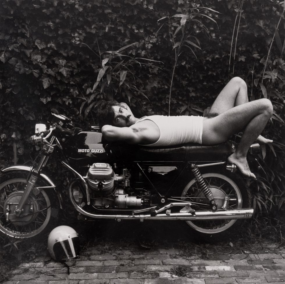 This is a black-and-white photograph by Robert Giard of a man in a white t-shirt with no pants reclining on a motorcycle.