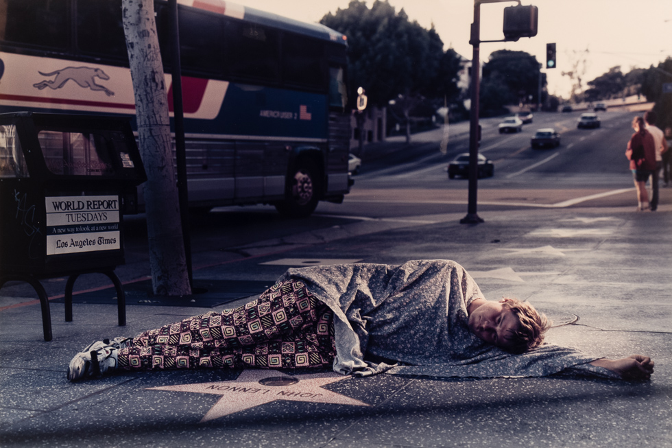 This is a color photograph of a man reclined on the ground with a blanket next to a Hollywood star on the sidewalk.