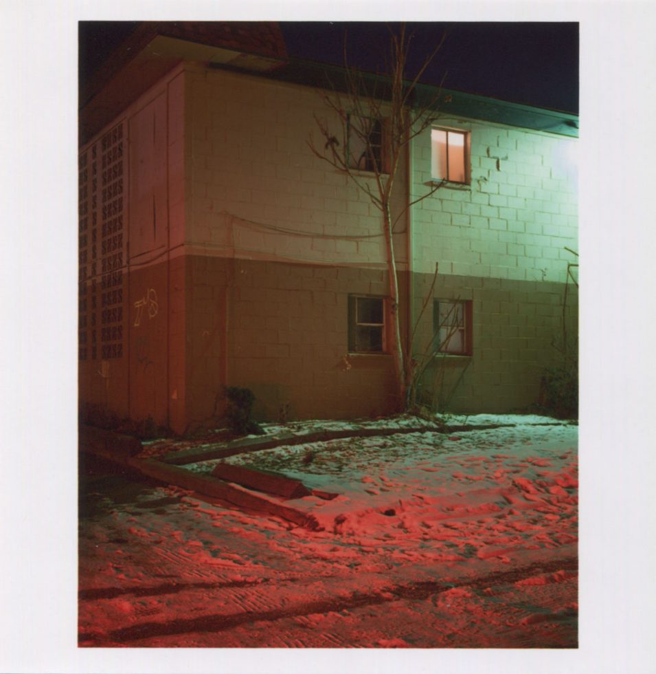 A color photograph by Todd Hido of the side of a house at night with green and red lights reflecting off the house and snowy foreground.