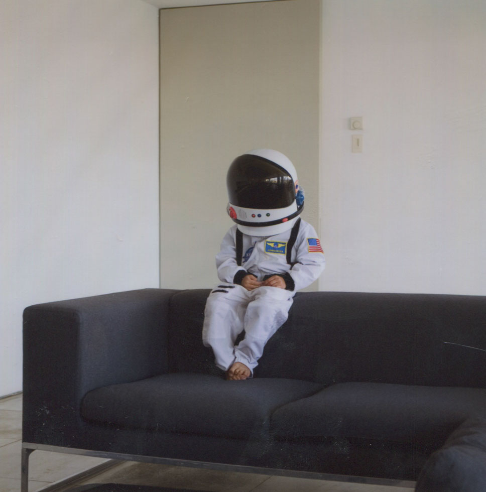 This is a color photograph of a child in a space suit sitting on a modern couch.