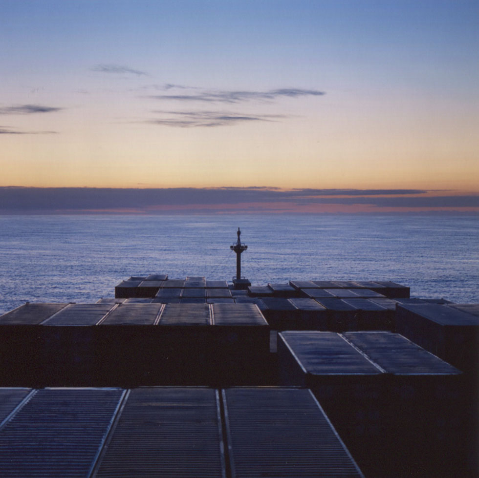 This is a photograph of containers on a boat sailing across a blue ocean with sunrise or sunset on the horizon.