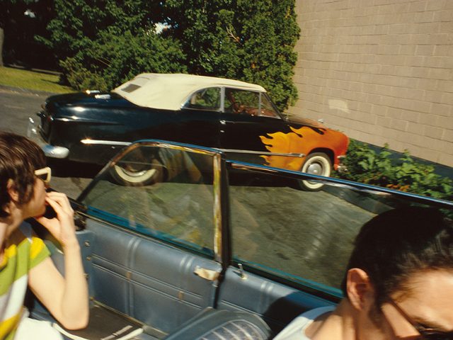 A 50s style convertible car, with flames painted on it photographed from another convertible car driving by with person in the backseat looking at the painted car