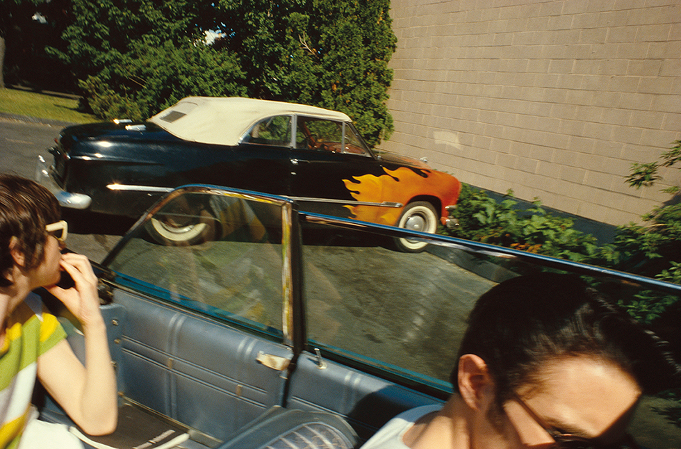 This is a color photo of a 50s-style convertible car with flames painted on it photographed from another convertible car driving by with person in the backseat looking at it.