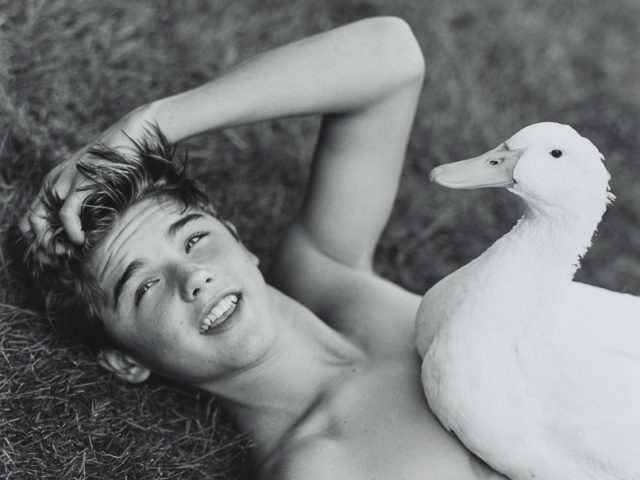 Photograph of a Young man lying in the grass with a duck sitting on his chest