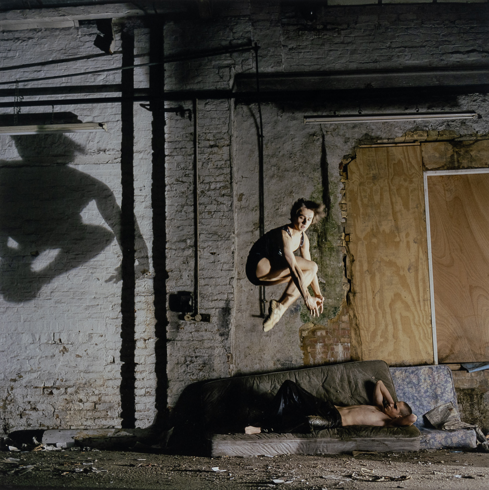 A color photograph of a man leaping above another man who is reclined on a mattress on the ground.