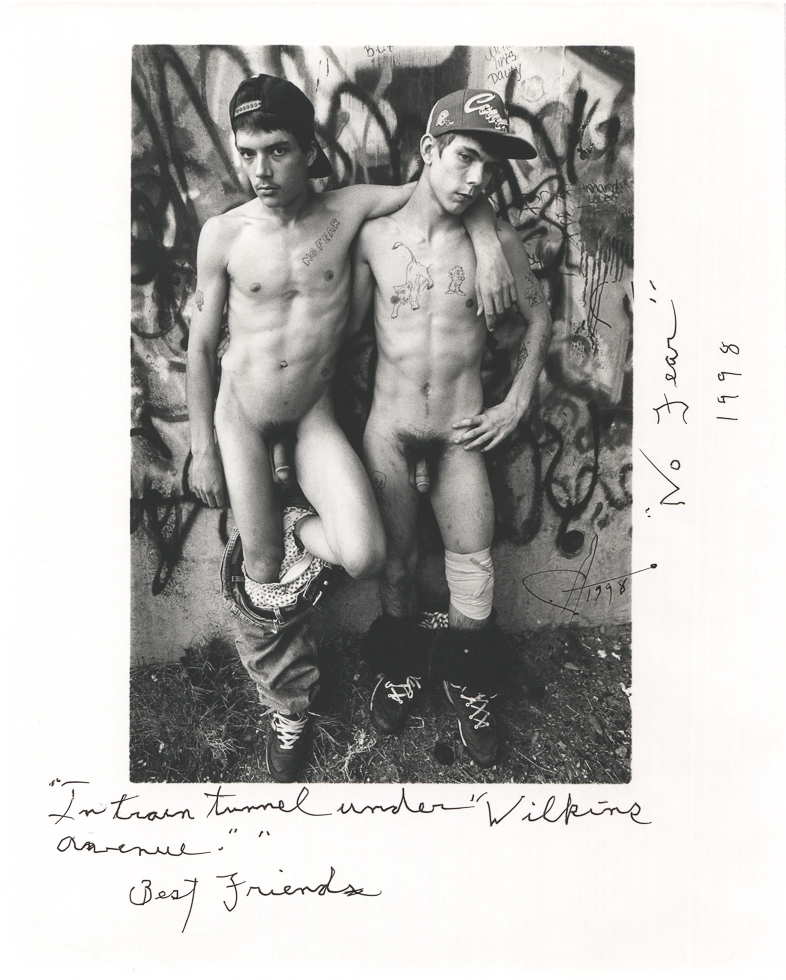 This is a black-and-white photograph of two naked men in hats against a graffiti wall with their arms around each other and handwriting around the image.