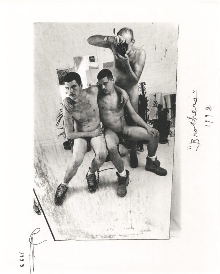 This black and white photograph shows three naked men sitting close to one another in a mirror.