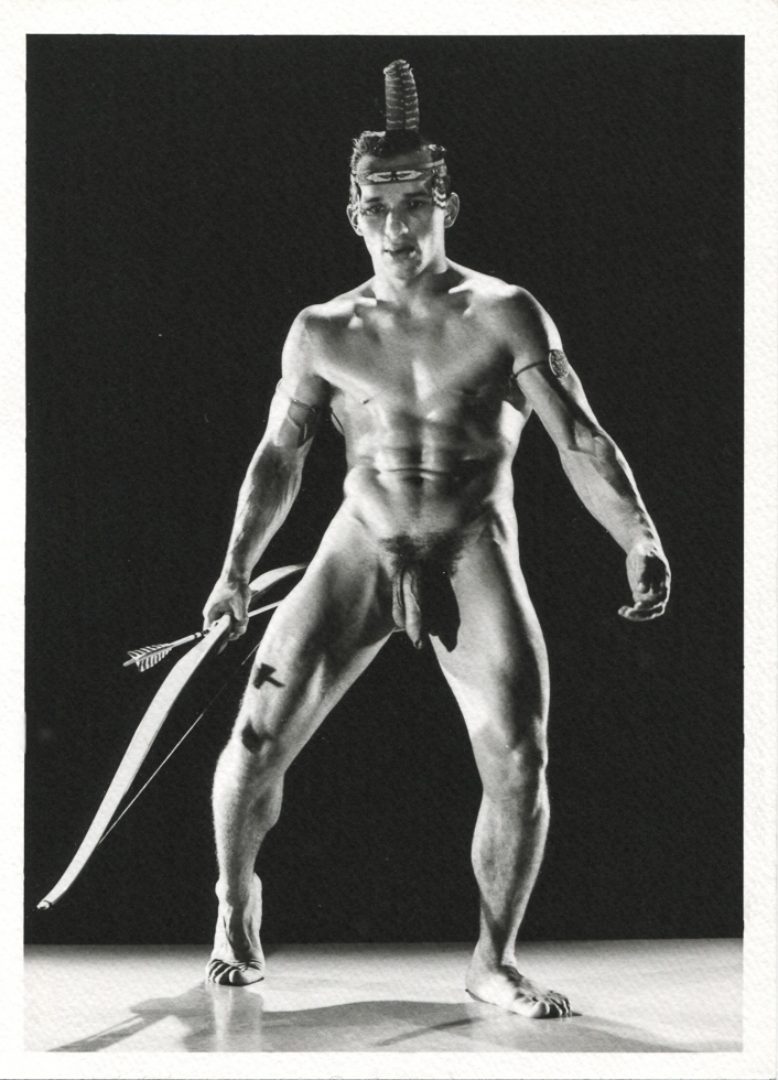 A black and white photograph on a nude man wearing Native American clothing holding a bow and arrow taken by Bruce Bellas, also known as Bruce of Los Angeles