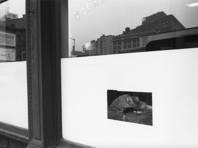 This is a black-and-white photograph of a restaurant window through which one can see a man asleep with his head face down on a table.