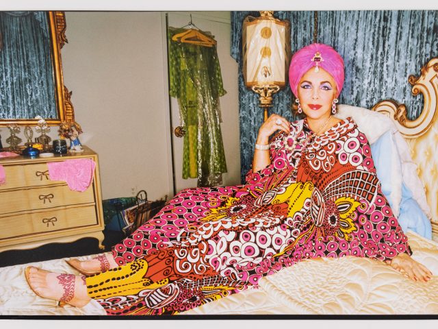 This is a color photograph of a woman in a pink kaftan lounging in a bed.