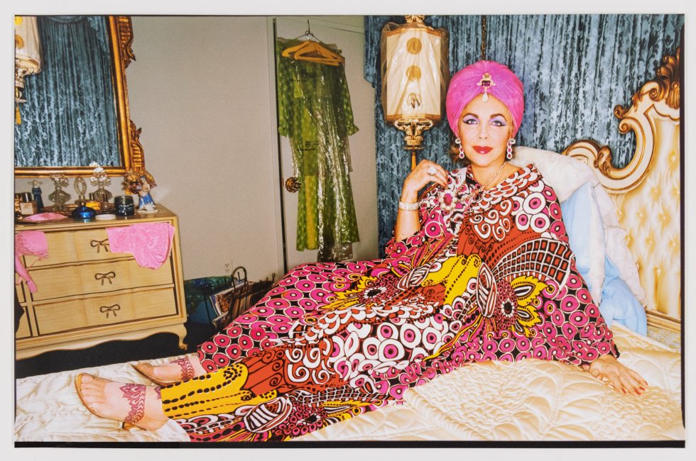 This is a color photograph of a woman in a pink kaftan lounging in a bed.