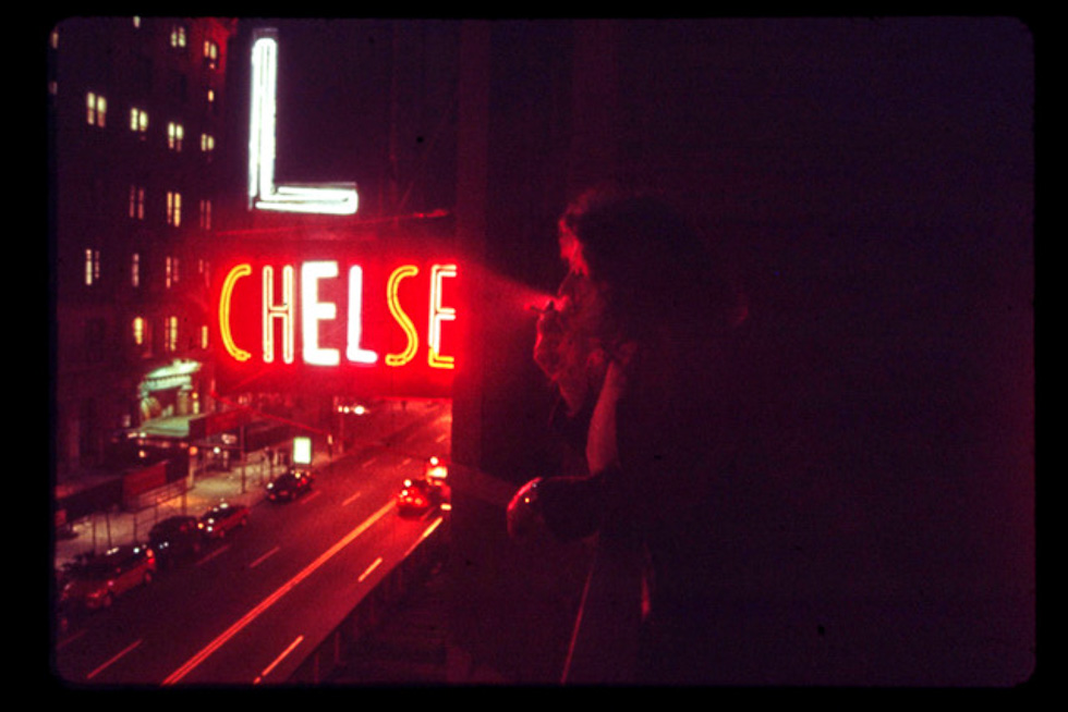 This is a color photograph of a person smoking a cigarette on the balcony of the Chelsea Hotel at night with red neon light.