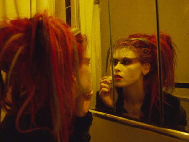 Linda Troeller made this yellow tone picture of a woman in club kid makeup and hair puts on makeup in a bathroom mirror in 1995 at the Chelsea Hotel.