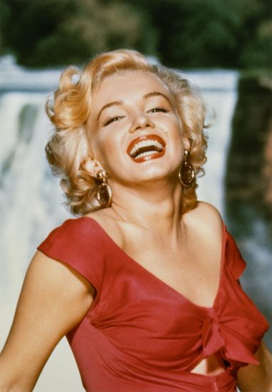 This is a color photograph of blonde woman laughing wearing a low-cut red dress.