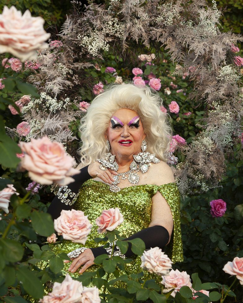 This is a photograph of the legendary drag performer Darcelle XV taken by Harry Hanson and Devin Antheus showing the icon against prink rose bushes wearing a glittery green dress.