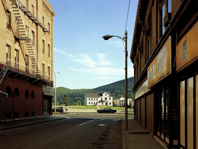 This is a photograph by Stephen Shore of Holden Street in the shade with two parked cars and a white house in the sun in the distance.