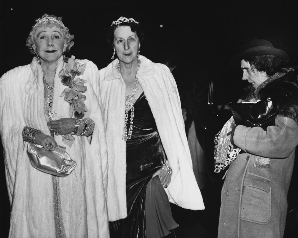 This is a black-and-white photograph of two women in furs and tiaras being scowled at by a homeless woman.