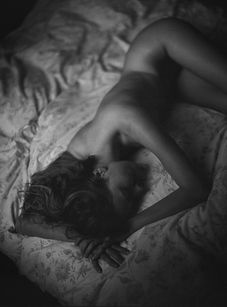 This is a black and white photograph of a nude woman lying in a bed taken from a birds eye view.