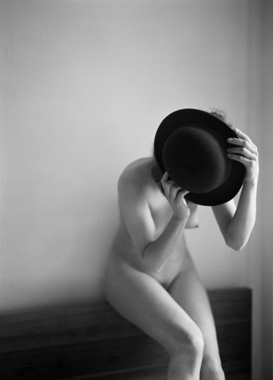 This is a black and white photograph of a nude woman holding a black hat over her face.