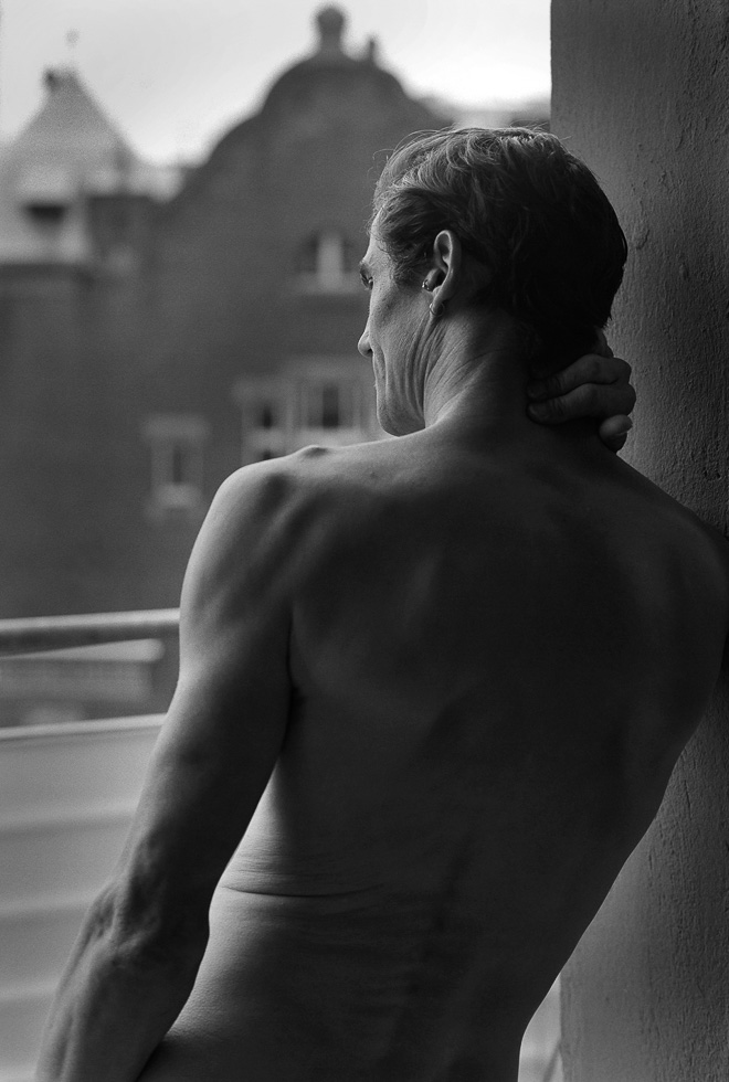 This is a black and white photograph of a nude man standing by a window.