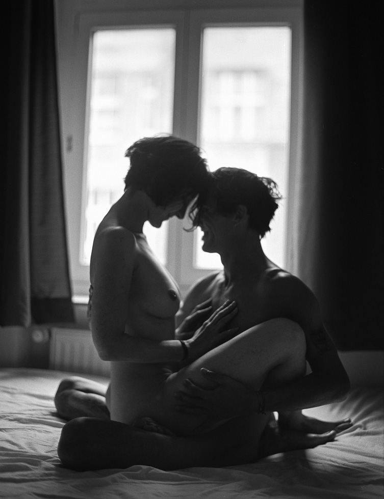 This is a black and white photograph of a nude couple embracing in a bed by a window.