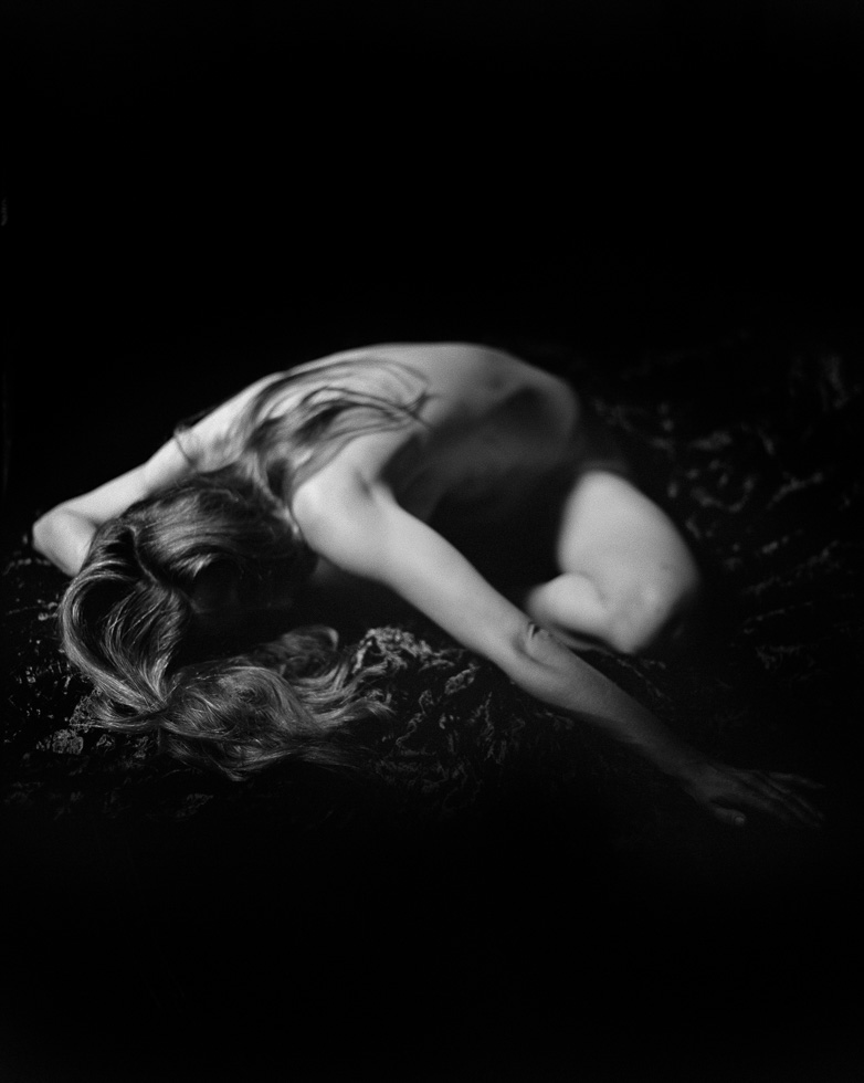 This is a black and white photograph of a nude woman seated on the ground with her head between her legs.