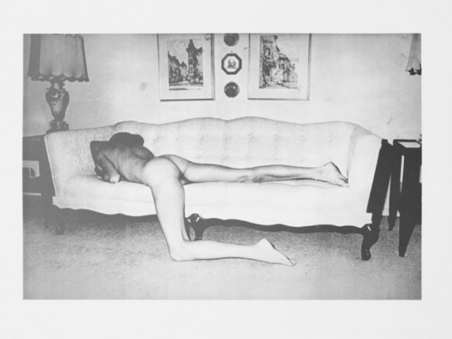 A black-and-white image of a nude men from behind stretched out on a light, tufted couch in a domestic setting.