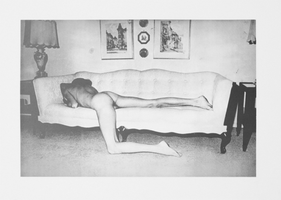 A black-and-white image of a nude men from behind stretched out on a light, tufted couch in a domestic setting.