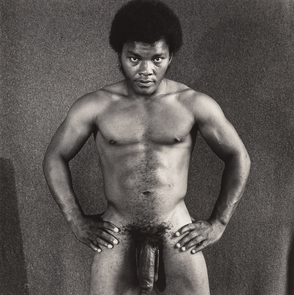 This is a black and white photograph of a naked Black man facing the camera straight on with his hands on his hips.