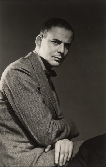 This is a black and white photograph of Lincoln Kirstein taken by George Platt Lynes circa 1940 with Lincoln sitting sideways in a blazer staring at the camera.