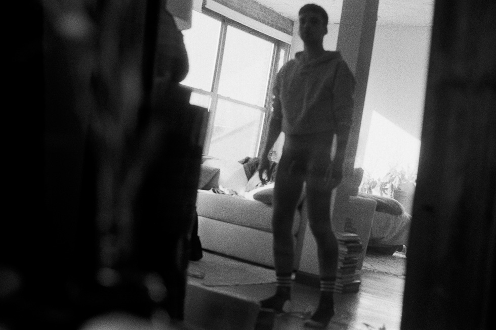 This is a black and white photograph by Jan Rattia of a boy in a sweatshirt, nude from the waist down standing in a living room.