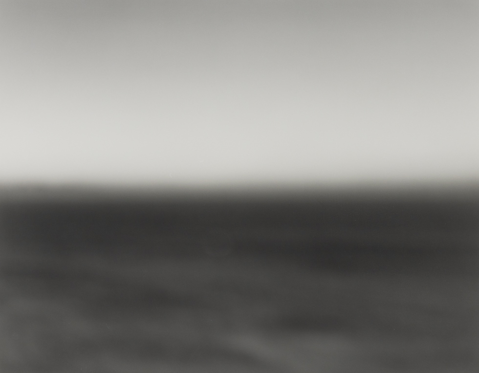 This is a blurry black and white photograph of the ocean that simply shows the horizon line of the dark sea below a blank grey sky.