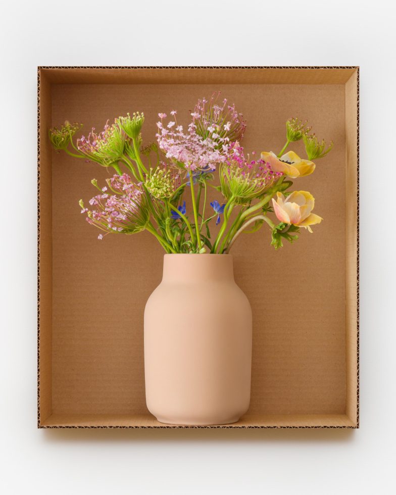 This is a photograph of flowers in a pink vase inside a cardboard box.