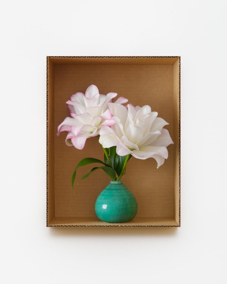 This is a photograph of flowers in a teal vase inside a cardboard box.