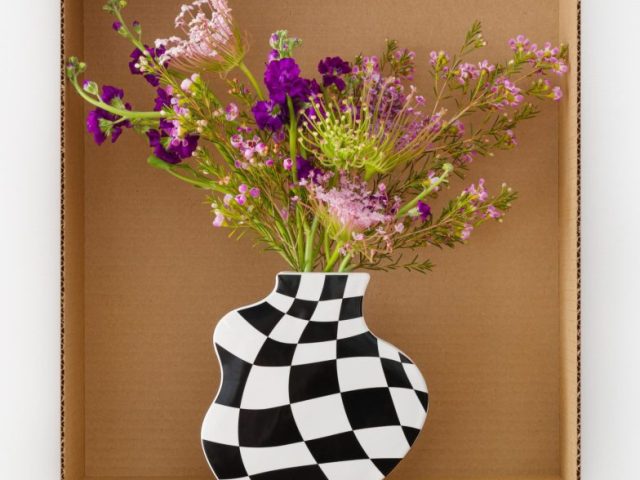 This is a photograph of purple flowers in a black and white checker vase inside a carboard box..