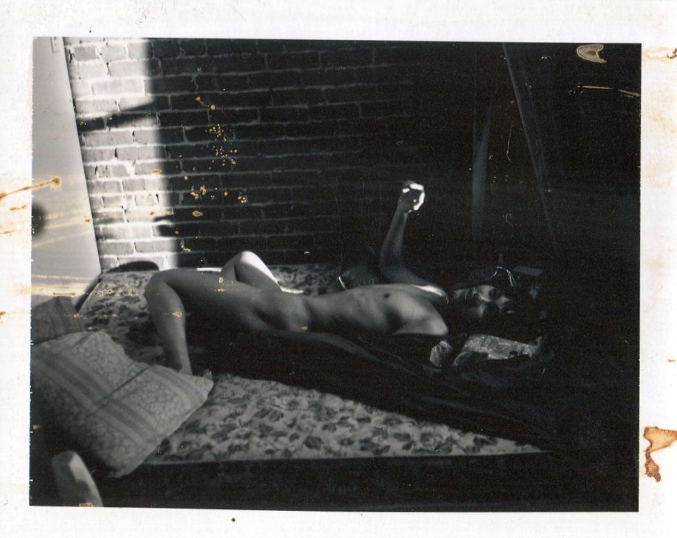 This is a black and white photograph of a naked person lying on a mattress next to a brick wall.